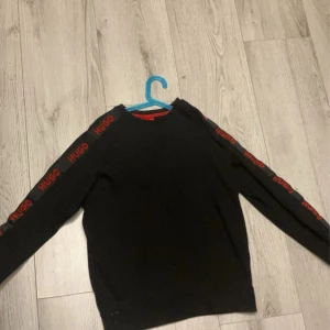 Svart tröja från Hugo - Snygg svart tröja från Hugo med röda logotyper längs ärmarna. Perfekt för en stilren look. Tröjan har långa ärmar och en klassisk passform. Ny skick, köpt på Room 1006 i Emporia. 