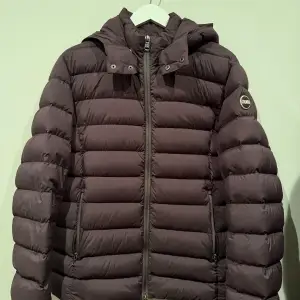 Säljer en brun dunjacka från Colmar med huva och dragkedja. Jackan har en quiltad design och är perfekt för kyligare väder. Den har en stilren look med en logotyp på ärmen. Färgen är coffee som drar något år t det vinröda hållet. Det finns en liten diskret lagning på ena armen, som ses på fjärde bilder. 