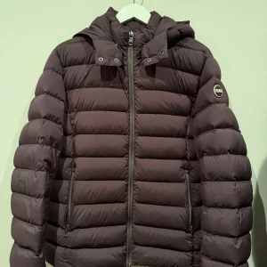 Brun dunjacka från Colmar - Säljer en brun dunjacka från Colmar med huva och dragkedja. Jackan har en quiltad design och är perfekt för kyligare väder. Den har en stilren look med en logotyp på ärmen. Färgen är coffee som drar något år t det vinröda hållet. Det finns en liten diskret lagning på ena armen, som ses på fjärde bilder. 