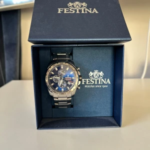 Silverfärgad klocka från Festina - Snygg och stilren klocka från Festina med en blå urtavla och silverfärgad länk. Klockan har flera sub-dials och datumvisning, perfekt för den som gillar detaljer. Levereras i originalförpackning. Tveka inte på att höra av er om det är något! Med vänlig hälsning, viktor