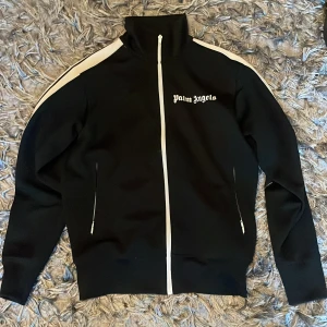 Svart track jacket från Palm Angels - Snygg svart track jacket från Palm Angels med vita ränder längs ärmarna och dragkedja framtill. Jackan har en hög krage och vita detaljer som ger en sportig look.