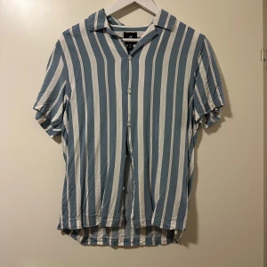Randig kortärmad skjorta från H&M - Snygg och avslappnad skjorta från H&M i blått och vitt randigt mönster. Skjortan har korta ärmar och knappar framtill, perfekt för en somrig look. Passar bra till både jeans och shorts.