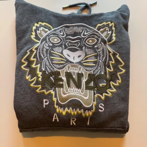 Grå hoodie från Kenzo med tigerbroderi - Säljer en snygg grå hoodie från Kenzo med ett stort, detaljerat tigerbroderi på framsidan. Hoodien har en justerbar huva med dragsko och ribbade muddar vid ärmslut och nederkant. Perfekt för en stilren och bekväm look.
