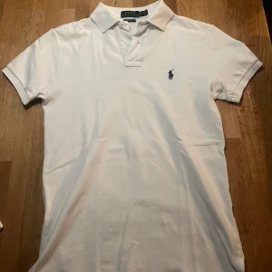 Vit pikétröja från Ralph Lauren - Snygg vit pikétröja från Ralph Lauren med klassisk krage och knappar. Den har en broderad logotyp i blått på bröstet och är i en custom fit passform. Perfekt för en stilren look. Pikén är i storlek XS och är i mycket bra skick