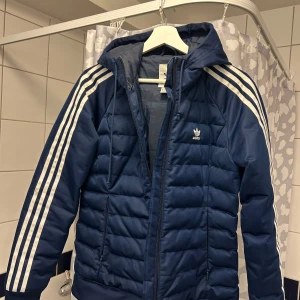 Blå dunjacka från Adidas - Säljer en snygg blå dunjacka från Adidas med de klassiska tre vita ränderna längs ärmarna. Jackan har en dragkedja framtill och en praktisk huva. Perfekt för kyligare dagar.