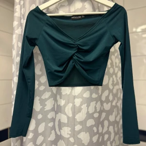 Mörkgrön croppad topp från PrettyLittleThing - Snygg mörkgrön croppad topp från PrettyLittleThing med v-ringning och rynkdetalj framtill. Toppen har långa ärmar och är perfekt för en trendig look.