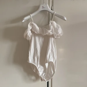 Vit offshoulder body från Zara - Säljer en söt vit offshoulder body från Zara med volangdetaljer. Perfekt för sommarens varma dagar. Den har smala axelband och en luftig passform.