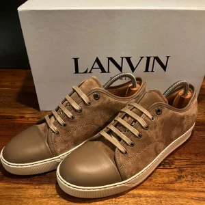 Bruna sneakers från Lanvin - Snygga bruna sneakers från Lanvin i mocka och skinn. Skorna har en klassisk design med snörning och en vit sula. Perfekta för en stilren look.