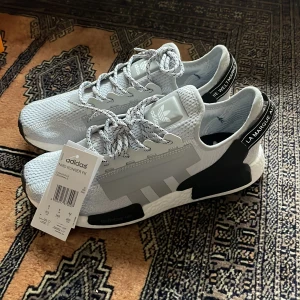 Grå Adidas NMD - Snygga grå Adidas NMD R1 sneakers med vit och svart sula. Skorna har en modern design med snörning och märkets logga på plösen. Perfekta för en stilren look. Helt nya oanvända✅