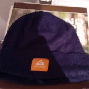 Mörkblå mössa från Garphyttan - Säljer en snygg mörkblå mössa från Garphyttan. Den är perfekt för kyliga dagar och har en stilren design med en liten orange etikett framtill. Mössan är mjuk och bekväm, idealisk för både vardag och utomhusaktiviteter.