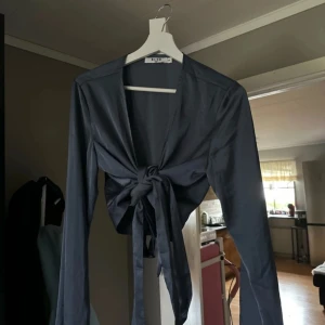 Mörkblå knytblus från NA-KD - Snygg mörkblå blus från NA-KD med knytning framtill. Perfekt för både vardag och fest! Den har långa ärmar och är gjord i ett mjukt material som känns skönt mot huden. Passar bra till jeans eller kjol för en stilren look. Aldrig använd.