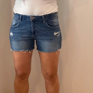 Blå jeansshorts med slitningar - Säljer ett par snygga blå jeansshorts med slitningar och fransiga kanter. De har en knapp och dragkedja fram samt fem fickor. Perfekta för sommaren och ger en avslappnad look. Passar till både vardag och fest!