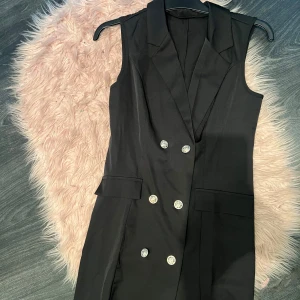 Svart ärmlös klänning med knappar - Snygg svart ärmlös klänning med dubbelknäppning och stiliga knappar. Perfekt för en elegant look, kanske till en fest eller ett speciellt tillfälle. Klänningen har en klassisk krage och är i en stilren design.