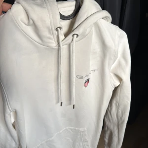 Vit hoodie från GANT - Säljer en snygg vit hoodie från GANT i storlek XS. Den har en klassisk design med GANT-loggan broderad på bröstet och en praktisk känguruficka. Perfekt för en avslappnad stil och passar till alla årstider. Huvan har justerbara snören för extra komfort.