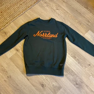 Mörkgrön och kamouflage tröja från SQRTN Company - Säljer två snygga tröjor från SQRTN Company i storlek XXS. Den första är mörkgrön med orange text 'The Great Norrland' på bröstet. Den andra har ett coolt kamouflagemönster med vit text 'Made In Norrland'. Båda tröjorna är långärmade och perfekta för höst och vinter. De är i bra skick och superbekväma!