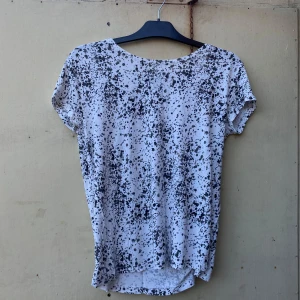 Vit och svart t-shirt från Saints and Mortals - Säljer en cool vit t-shirt från Saints and Mortals med ett svart stänkmönster. Den har korta ärmar och en avslappnad passform, perfekt för en casual look. Materialet känns mjukt och skönt mot huden.