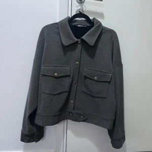 Grå jacka från Gina Tricot - En snygg grå jacka från Gina Tricot i storlek XL. Jackan har en klassisk krage och knappar framtill. Den har två stora fickor på bröstet och långa ärmar med knappar vid ärmsluten. Perfekt för höst och vår!