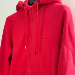 Röd hoodie från Peak Performance - Säljer en snygg och bekväm röd hoodie från Peak Performance. Den har en dragkedja framtill och justerbar huva med snören. Nypris ligger på ungefär 1200 kronor men jag säljer för 650 kronor. Den är i bra skick och det enda som saknas är ett av snörernas svarta ända. Som man kan se på bilden har den åkt av annars inga defekter. 