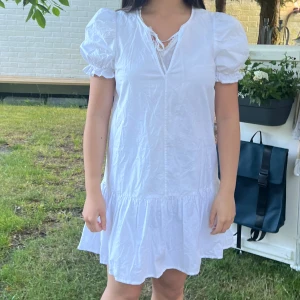 Vit kortklänning med puffärmar - Säljer en superfin vit kortklänning med puffärmar och volangdetaljer. Klänningen har en v-ringad hals med snörning och är perfekt för sommaren. Den är gjord av ett lätt och bekvämt material, perfekt för varma dagar. Passar bra till både vardag och fest!