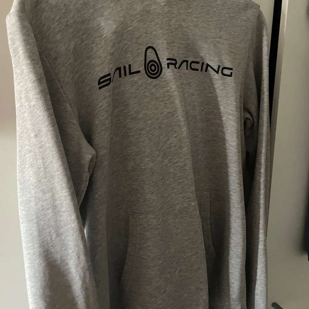 Säljer en grå hoodie från Sail Racing. Den är superbekväm och perfekt för kyliga dagar. Hoodien har en stor ficka på magen och en snygg logga på bröstet. Den är i bra skick och passar perfekt för både vardag och träning. Köp den nu eller ge ett bud! 💛. Hoodies.