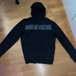Under Armour windbreaker  - Säljer en svart windbreaker från Under Armour med dragkedja och huva. Den har en grå detalj över bröstet med logotyp. Perfekt för en avslappnad stil. Storlek S. 