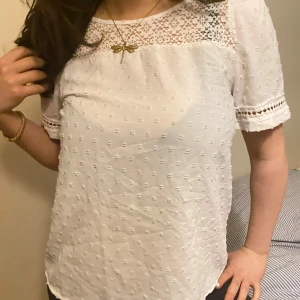 Vit blus med spetsdetaljer - Säljer en söt vit blus med spetsdetaljer på axlarna och ärmarna. Blusen har korta ärmar och ett diskret prickigt mönster. Perfekt för en somrig look!