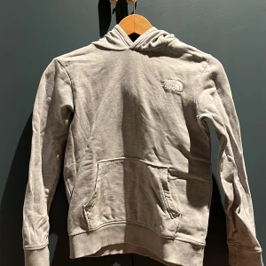 Grå hoodie från The North Face - Säljer en stilren grå hoodie från The North Face. Den har en klassisk design med en känguruficka framtill och en liten logga på bröstet. Perfekt för en avslappnad look.Lite lagad som man ser på den fjärde bilden men det syns knappt