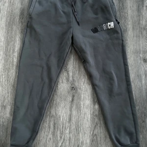 Grå mjukisbyxor från Hoodrich - Snygga grå mjukisbyxor från Hoodrich med broderad logga i svart och vitt. Byxorna har en bekväm passform med snörning i midjan och ribbade muddar vid bensluten. Perfekta för en avslappnad stil.