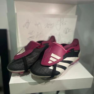 Adidas Predator fotbollsskor - Snygga Adidas Predator fotbollsskor i svart och vinrött med vita detaljer. Skorna har en unik design med snörning och en skyddande flik över snörningen. Yttersulan är mönstrad för optimalt grepp på planen.