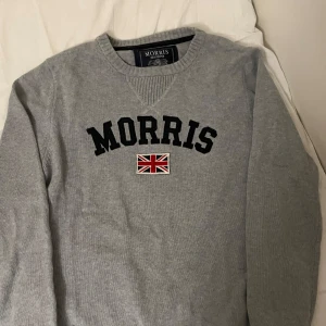 Grå tröja från Morris - Säljer en stilren grå tröja från Morris med mörkblå text och en brittisk flagga på framsidan. Tröjan har långa ärmar och en rund halsringning. Perfekt för en avslappnad och snygg look.