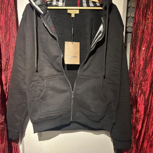 Svart kofta från Burberry - Snygg svart huvtröja från Burberry med dragkedja och klassiskt rutigt mönster i huvan. Tröjan har en broderad logga på bröstet och praktiska fickor framtill. Perfekt för en stilren look. OBS! Priset är inte hugget i sten. Hör av er vid minsta fundering 🍾