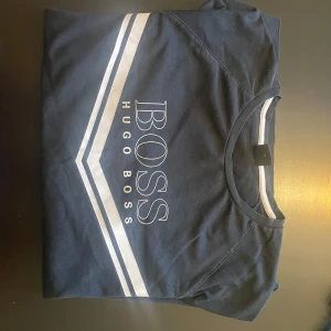 Svart sweatshirt från Hugo Boss - Säljer en s svart sweatshirt från Hugo Boss med vit text och ränder över bröstet. Tröjan har rund hals och långa ärmar, perfekt för en avslappnad look. Passar bra till både jeans och joggers.