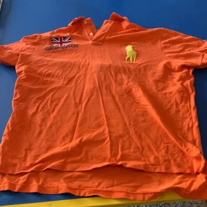 Orange pikétröja från Ralph Lauren ”chief keef polo” - Säljer en orange pikétröja från Ralph Lauren med broderad text 'Great Britain' och en brittisk flagga. Tröjan har en gul broderad siffra '3' på ryggen och en gul häst på framsidan. Perfekt för dig som gillar färgstarka plagg!