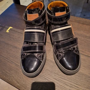 Svarta sneakers från Bally - Köpta för 5000 kronor. Snygga svarta sneakers från Bally med kardborreband och vita detaljer. Skorna har en hög modell med en brun insida och en robust sula. Perfekta för en stilren look. Den ena är en halv storlek större än den andra, märks inte med sula i.