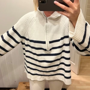 Vit och marinblå randig stickad tröja - Säljer en snygg vit stickad tröja med marinblå ränder och halv dragkedja. Tröjan har långa ärmar och en bekväm passform, perfekt för kyligare dagar. Den ribbade strukturen ger en extra touch av stil. Perfekt för en avslappnad look.