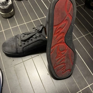 Grå sneakers från Christian Louboutin - Snygga svarta sneakers från Christian Louboutin med klassisk röd sula. Skorna har snörning och är tillverkade i mocka för en lyxig känsla. Perfekta för dig som vill ha stil och komfort i ett.