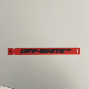 Rött armband från Off-White - Säljer ett coolt rött armband från Off-White med svart text. Armbandet har en minimalistisk design och är perfekt för att ge en edgy touch till din outfit. Det är tillverkat i ett slitstarkt material och har en enkel låsmekanism.