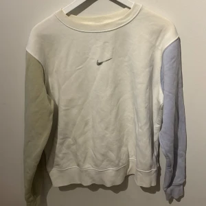 Vit sweatshirt från Nike - Säljer en stilren vit sweatshirt från Nike med en liten logga på bröstet. Tröjan har långa ärmar i lila och grön pastellfärg, vilket ger en unik look. Perfekt för en avslappnad stil.