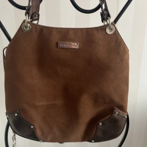 Brun axelväska från Betty Barclay - Snygg brun axelväska från Betty Barclay med detaljer i mörkare brunt och silverfärgade nitar. Väskan har en praktisk dragkedja och magnetknapp för säker förvaring. Perfekt för att bära dina dagliga nödvändigheter med stil.