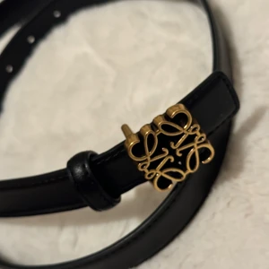 Svart bälte med gulddetalj - Snyggt svart bälte med en elegant guldfärgad spänne i form av en dekorativ design. Perfekt för att ge en stilren touch till din outfit. Tillverkat i ett material som liknar skinn.