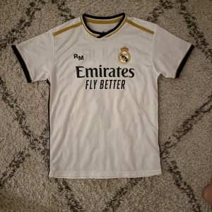 Vit Real Madrid fotbollströja - Säljer en vit Real Madrid fotbollströja med korta ärmar och svarta detaljer. Tröjan har klubbens emblem på bröstet och sponsorlogga 'Emirates Fly Better'. På baksidan finns namnet 'VINI JR.' och nummer 7. Perfekt för fans av laget! Priset går och diskutera vid en snabb affär!! Mvh Liam!! 