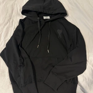 Svart hoodie från Ami - Säljer en stilren svart hoodie från Ami med en liten logga på bröstet. Tröjan har en klassisk design med huva och dragsko samt en praktisk magficka. Perfekt för en avslappnad look.