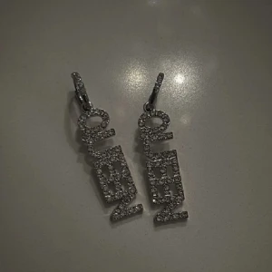Glittriga örhängen med texten 'QUEEN' - Snygga örhängen med texten 'QUEEN' i glittrande stenar. Perfekta för att ge en extra touch av glamour till din outfit. De hängande bokstäverna är täckta av små, gnistrande stenar som fångar ljuset vackert.