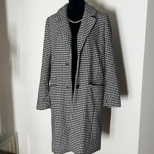 Svartvit rutig kappa - Snygg svartvit rutig kappa med klassiskt mönster och knappar framtill. Perfekt för att ge en stilren touch till din outfit. Kappan har en elegant krage och långa ärmar.