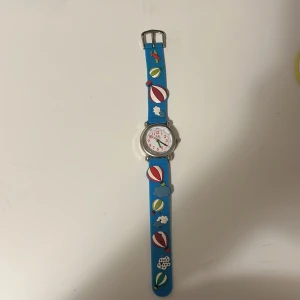 Blå barn klocka men luftballong motiv  . - Charmig barnklocka med blått armband dekorerat med färgglada luftballonger och moln. Klockan har en tydlig vit urtavla med röda siffror och visare. Perfekt för barn som lär sig klockan!
