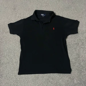 Svart pikétröja från Ralph Lauren - Säljer en klassisk svart pikétröja från Ralph Lauren med röd broderad logga på bröstet. Tröjan har korta ärmar och en stilren krage. Perfekt för en avslappnad men ändå stilfull look.