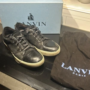 Svarta sneakers från Lanvin - Snygga svarta sneakers från Lanvin med vit sula och klassisk snörning. Skorna kommer med originalförpackning och tygpåsar för förvaring. Perfekta för en stilren look. Billigare vid snabb och smidig affär 