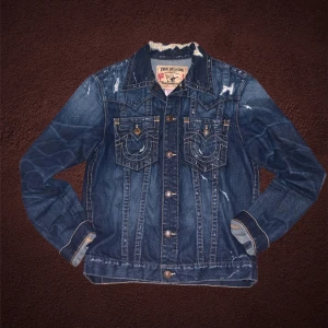 Mörkblå jeansjacka från True Religion - TAR ÄVEN BYTEN🦅🔥Snygg mörkblå jeansjacka från True Religion med slitna detaljer och knappar framtill. Jackan har två bröstfickor med lock och knapp samt en klassisk krage. Perfekt för en cool och avslappnad stil.