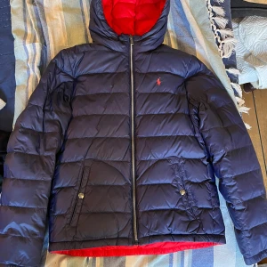 Blå och röd dunjacka från Ralph Lauren - Säljer en snygg blå och röd dunjacka från Ralph Lauren. Jackan har en dragkedja framtill och är quiltad för extra värme. Den har långa ärmar och en huva för att skydda mot kyla. Perfekt för vintern! Från djur/rökfritt hem!  Obs: Fri frakt! 