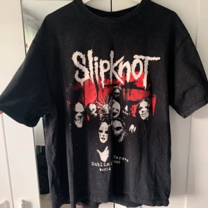 Svart Slipknot t-shirt från Bershka - Säljer en svart t-shirt från Bershka med Slipknot-tryck i vitt och rött. T-shirten har korta ärmar och ett coolt bandmotiv på framsidan. Perfekt för fans av bandet!